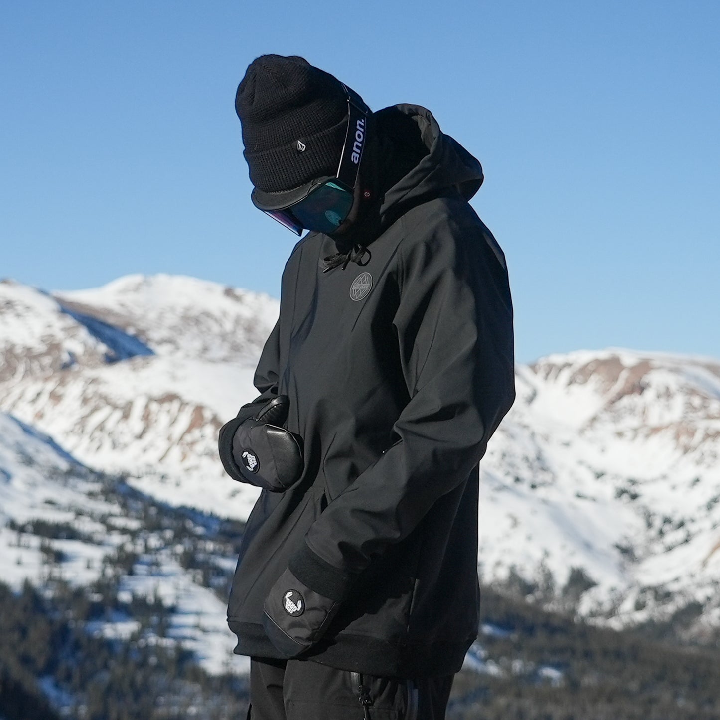 A person wearing a black waterproof riding hoodie with a hood, standing against a snowy mountain background. The hoodie has a kangaroo pocket, mesh pit vents, and zippers.