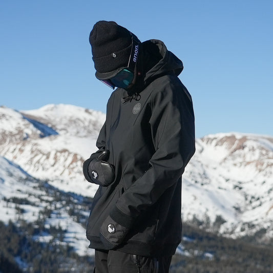 A person wearing a black waterproof riding hoodie with a hood, standing against a snowy mountain background. The hoodie has a kangaroo pocket, mesh pit vents, and zippers.